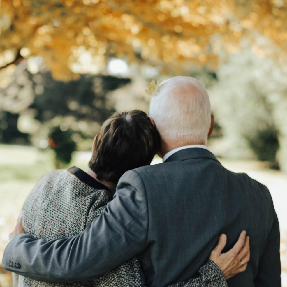 An older couple embraces, gazing at a fall-colored tree, outdoors.