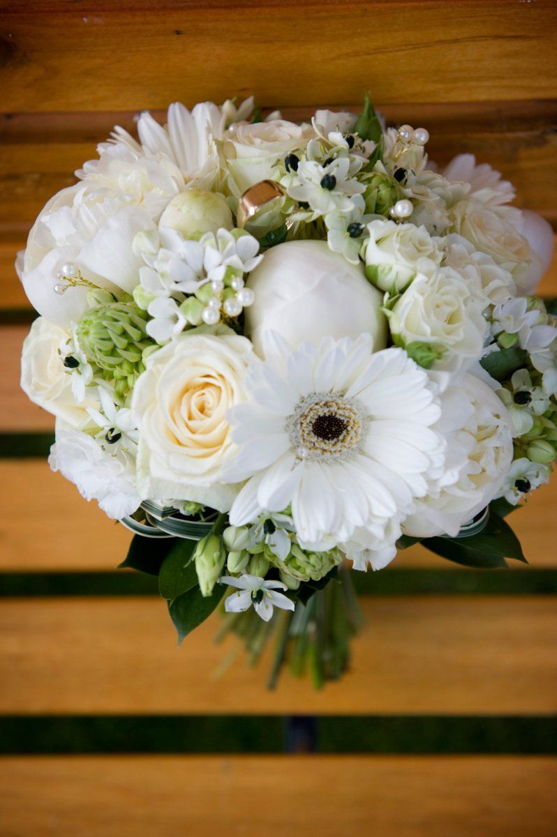 White floral bouquet on a wooden surface, featuring roses, peonies, and other white blooms.
