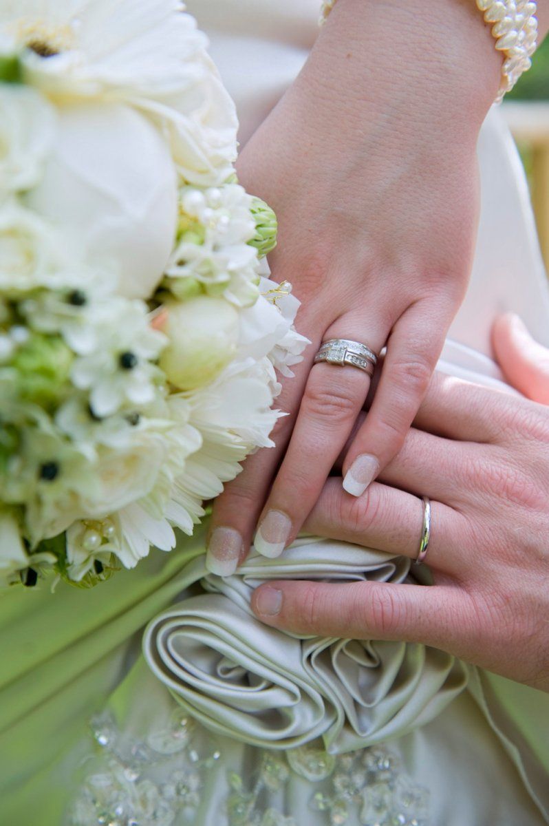 Hands with wedding rings on a white dress, holding a white flower bouquet.