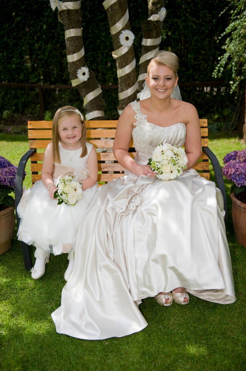 Bride in satin gown with flower girl, seated on a bench, holding bouquets, in a garden.