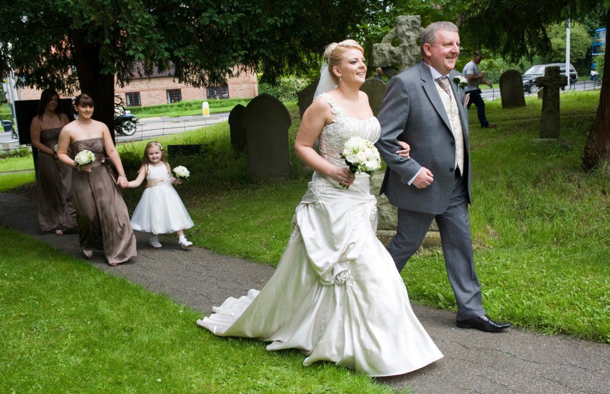 Bride and her father walk toward a church, bridesmaids and flower girl follow.