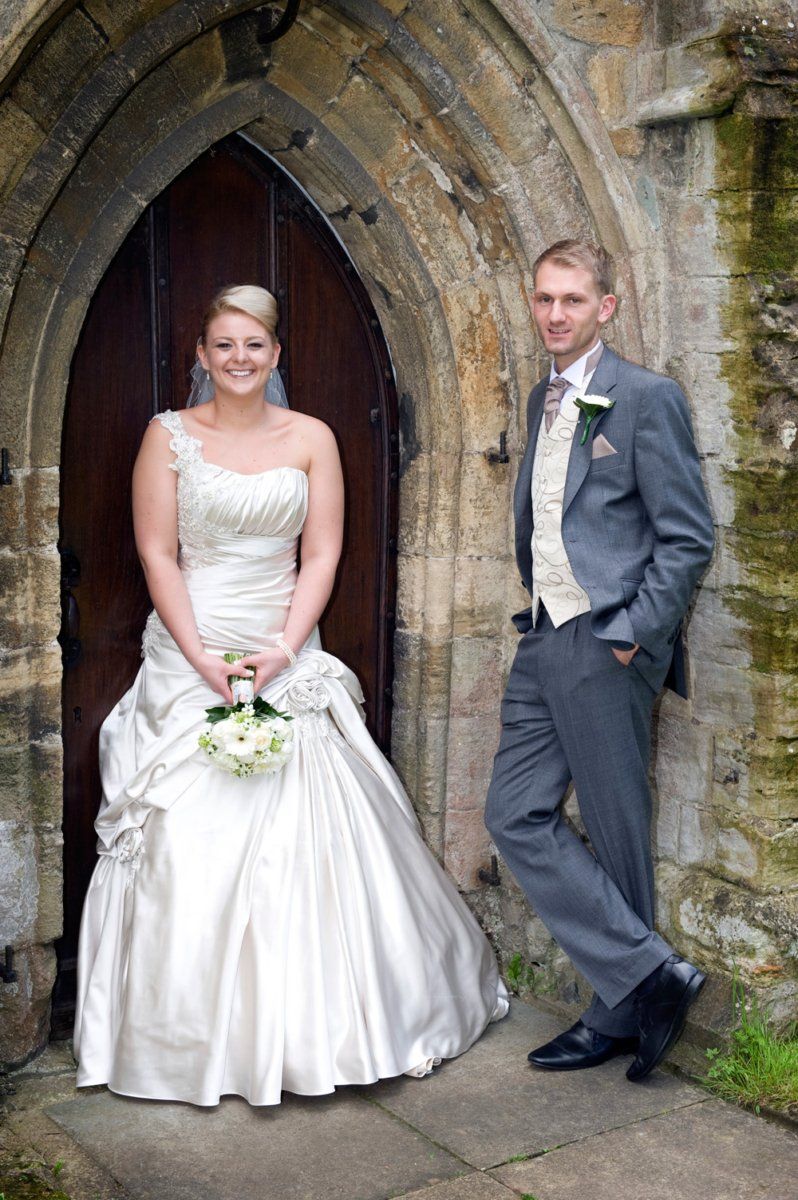 Bride and groom pose near a church door. The bride wears a white dress, holding a bouquet. The groom wears a gray suit.