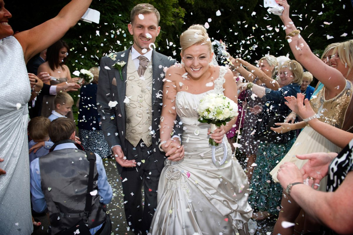 Newlyweds walk through confetti, smiling, with guests cheering.