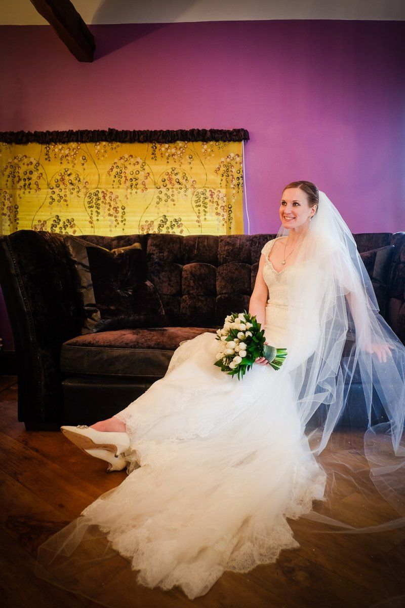 Bride in a white gown and veil sits on a dark brown couch, holding flowers, against a purple wall.