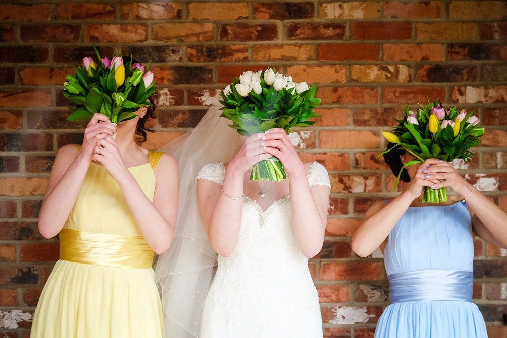 Bride and bridesmaids hiding faces behind bouquets of flowers in front of a brick wall.