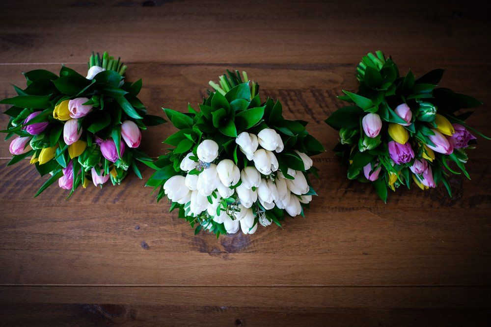 Three tulip bouquets on a wooden surface: one white, two with pink, yellow, and purple tulips, all with green leaves.