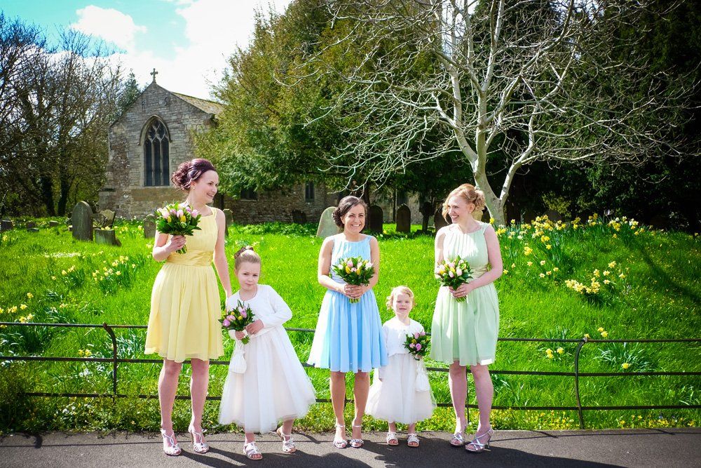 Bridesmaids in pastel dresses with bouquets pose near a church on a sunny day.