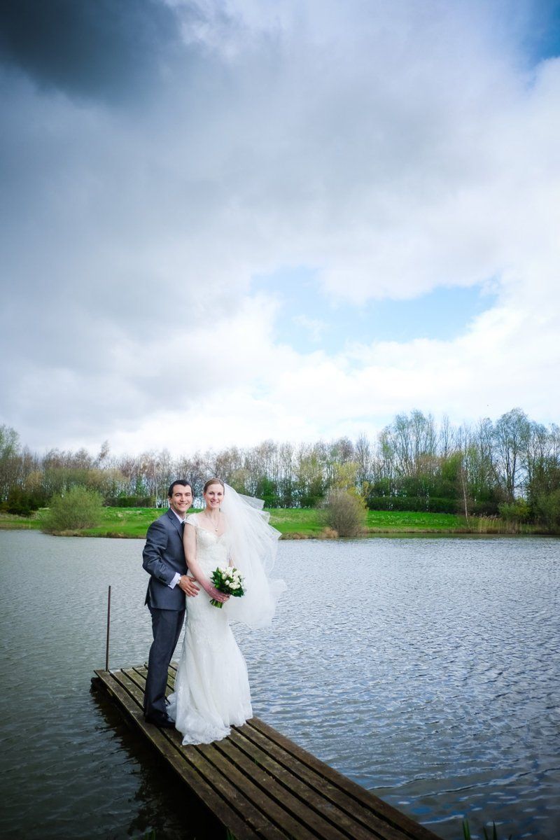 Bride and groom stand on a wooden dock over a lake, cloudy sky overhead.