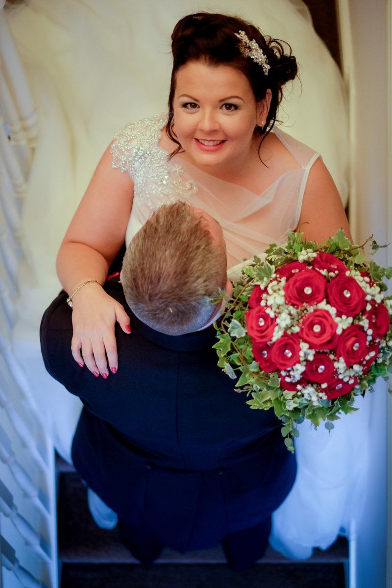 Bride smiles holding red rose bouquet as groom embraces her on stairs.