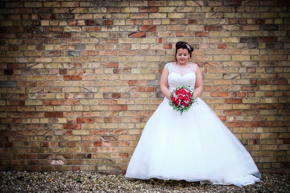 Bride in a white gown stands against a brick wall, holding a red bouquet.