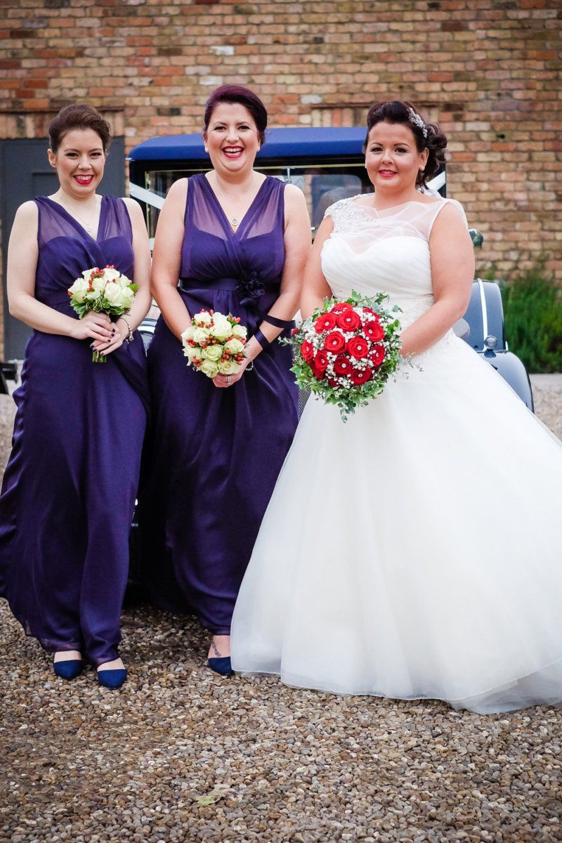 Bride with two bridesmaids posing near a vintage car; all are smiling.