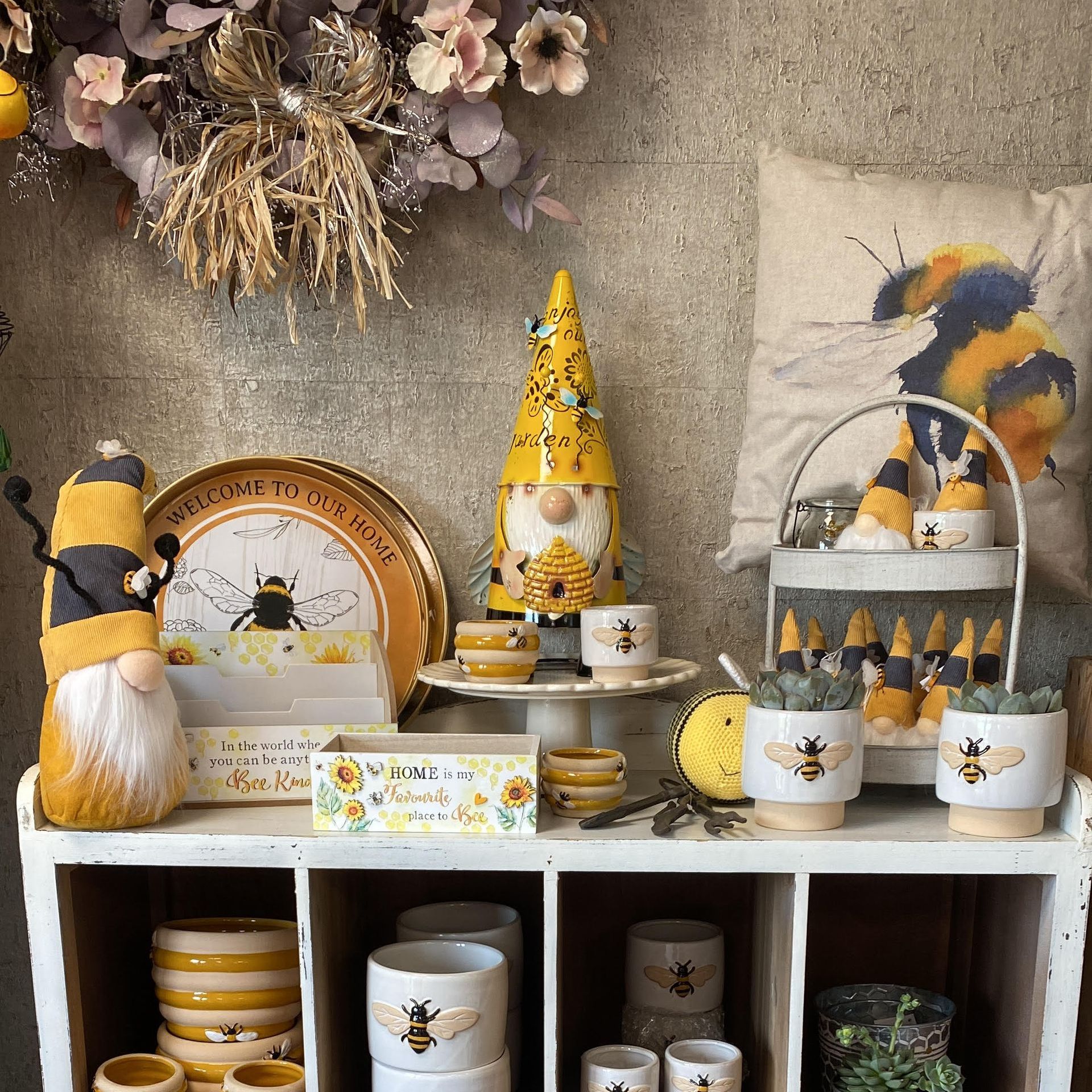 Display of bee-themed home decor on a white shelf. Includes gnomes, planters, and a pillow. Yellow and white colors.