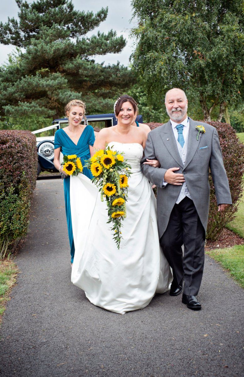 Bride, bridesmaid, and man walk down a path. Bride in white dress, holding sunflowers; bridesmaid in teal dress. Man in suit.