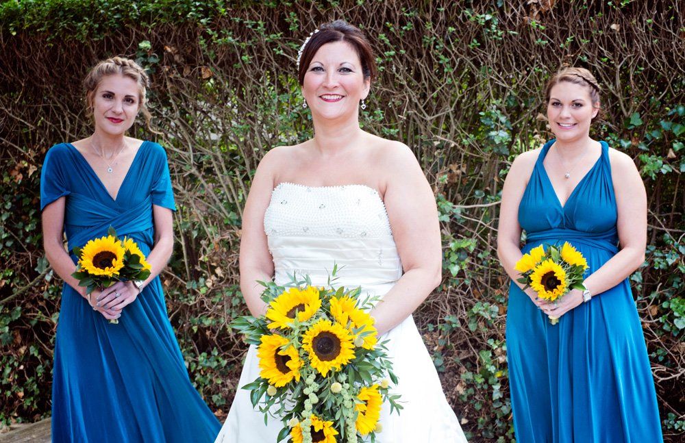 Bride with two bridesmaids holding sunflowers. All are smiling, wearing dresses.