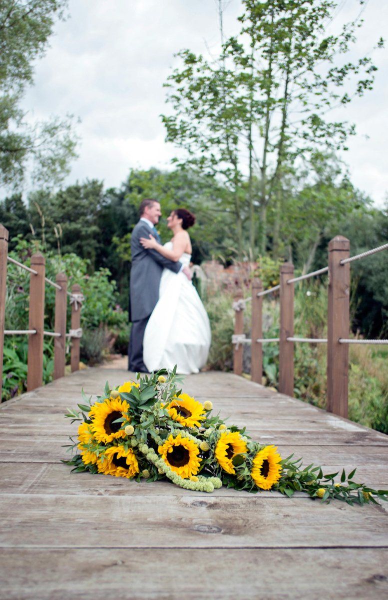 Wedding couple embraces on a wooden bridge, sunflowers in foreground, surrounded by greenery.
