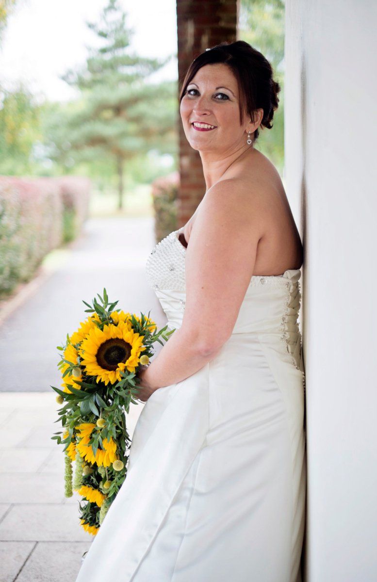 Bride in strapless white dress, smiling, holding sunflower bouquet, leaning against wall outdoors.