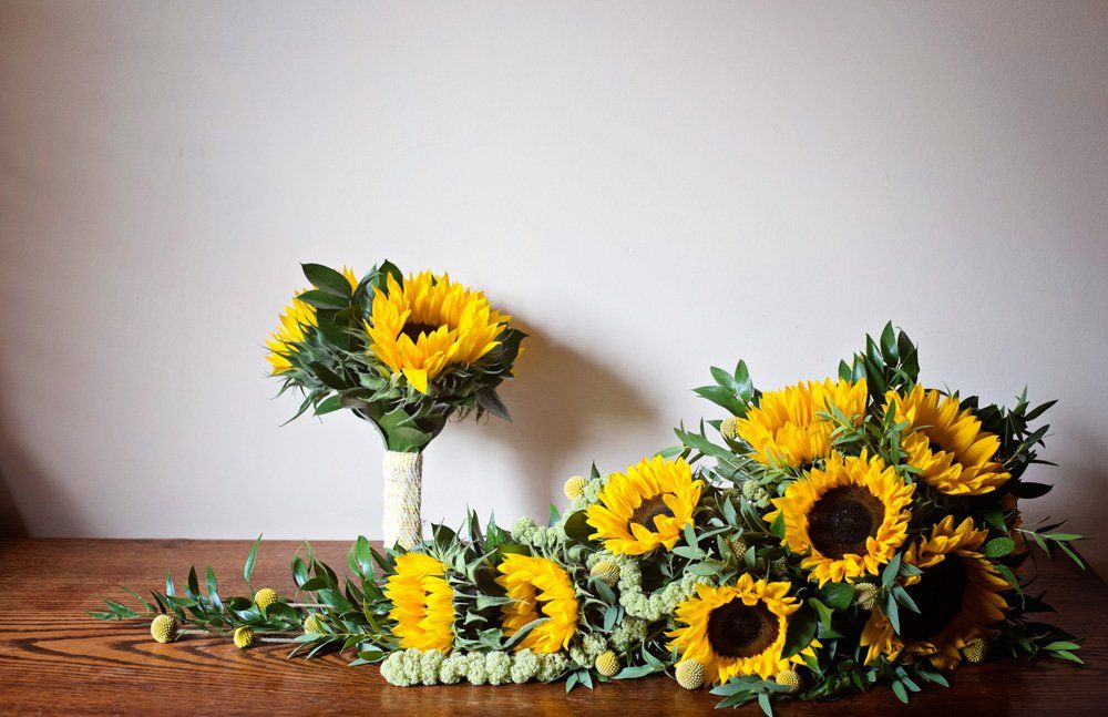 Sunflowers bouquets on a wooden surface, one small, one trailing; against a neutral wall.