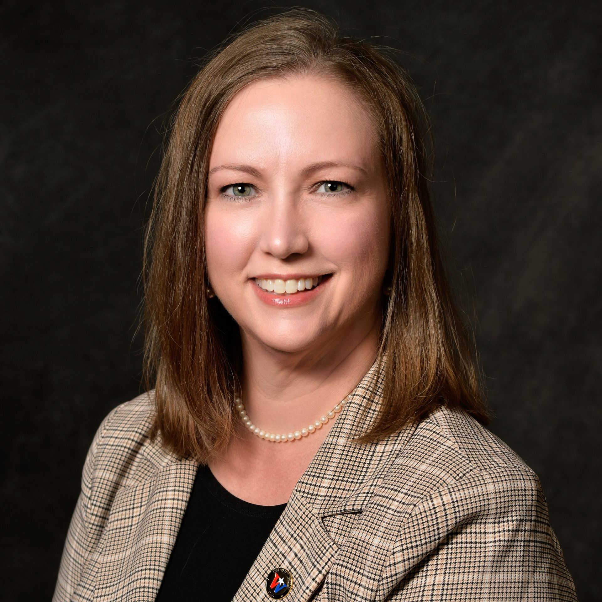 A woman wearing a plaid jacket and a pearl necklace is smiling for the camera.