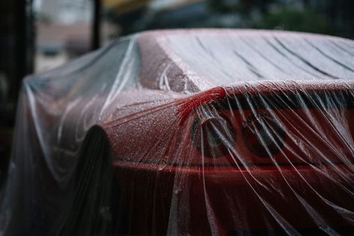Red car covered in a clear tarp, beaded with water droplets, parked outdoors.