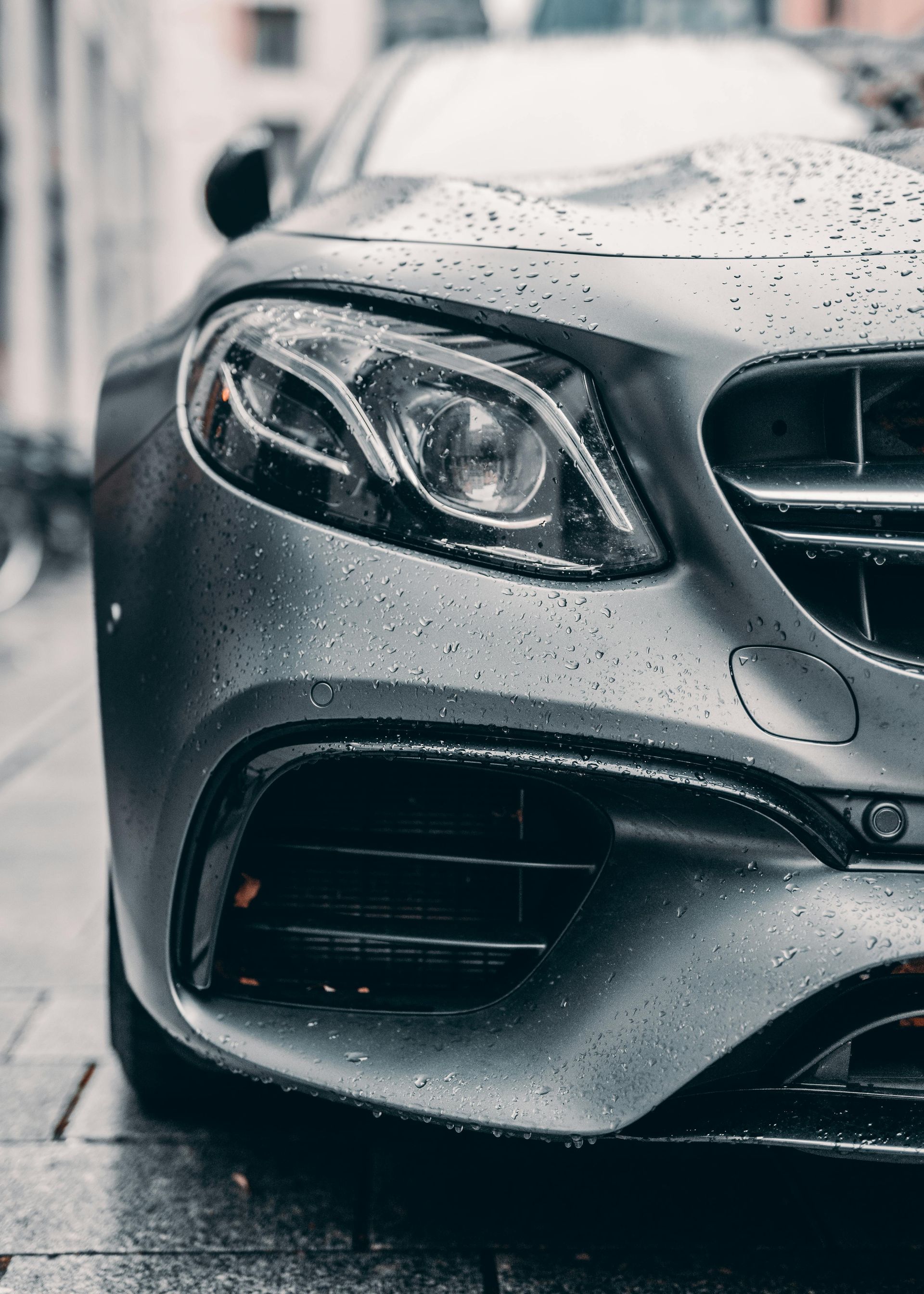Gray Mercedes-Benz car parked on a wet cobblestone street; raindrops on the hood and headlights.
