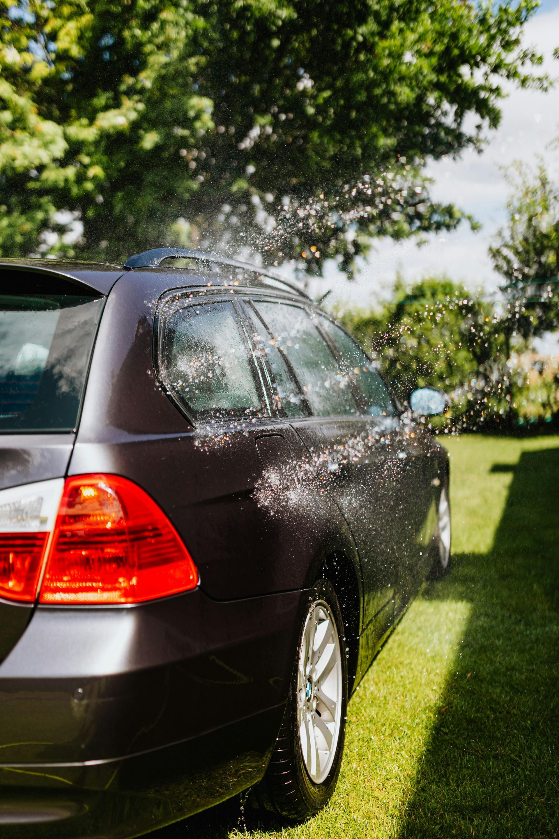 Car being washed with water spray on a sunny day in a grassy yard.