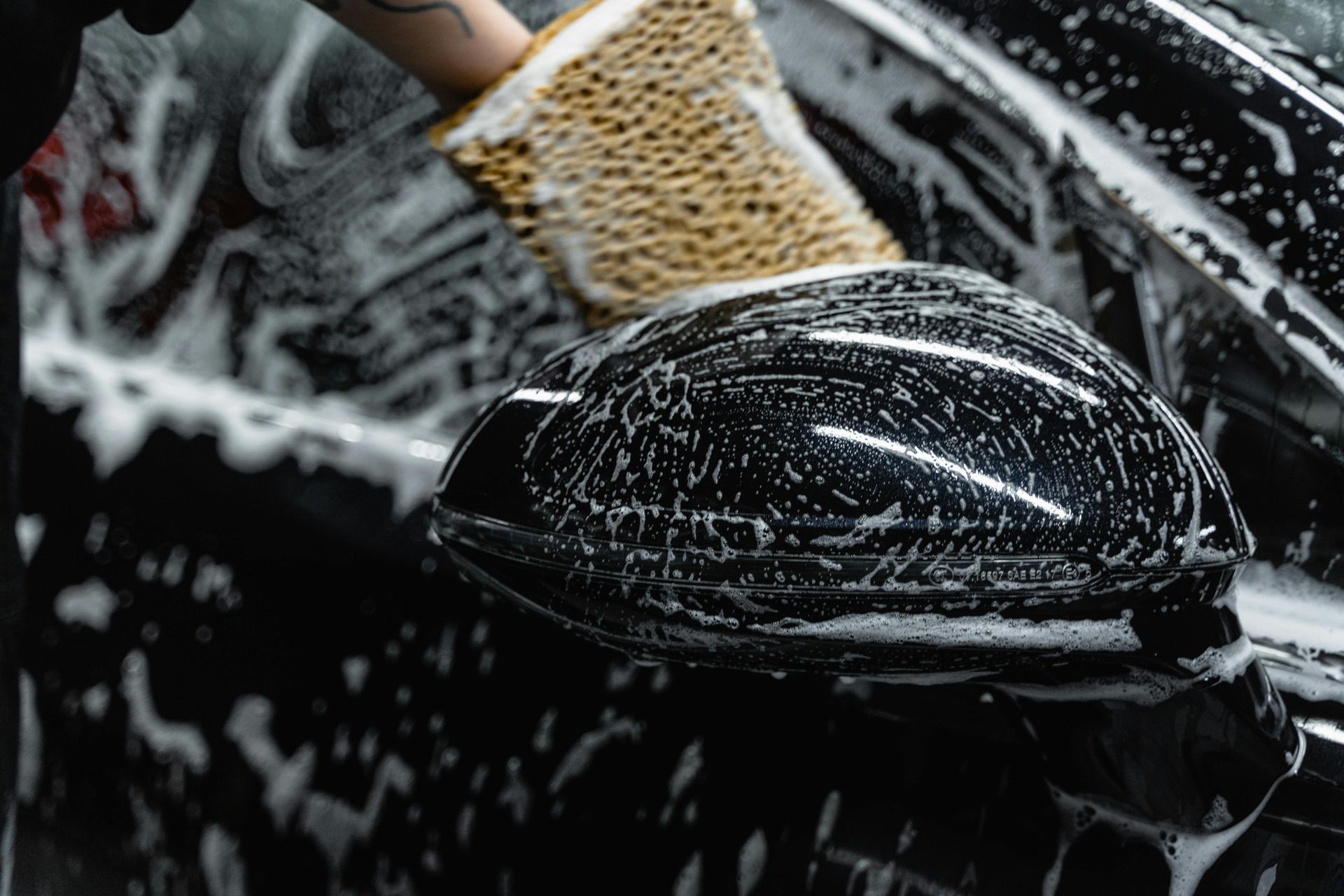 Person washing a black car with soapy water and a sponge. Focus on the side mirror.