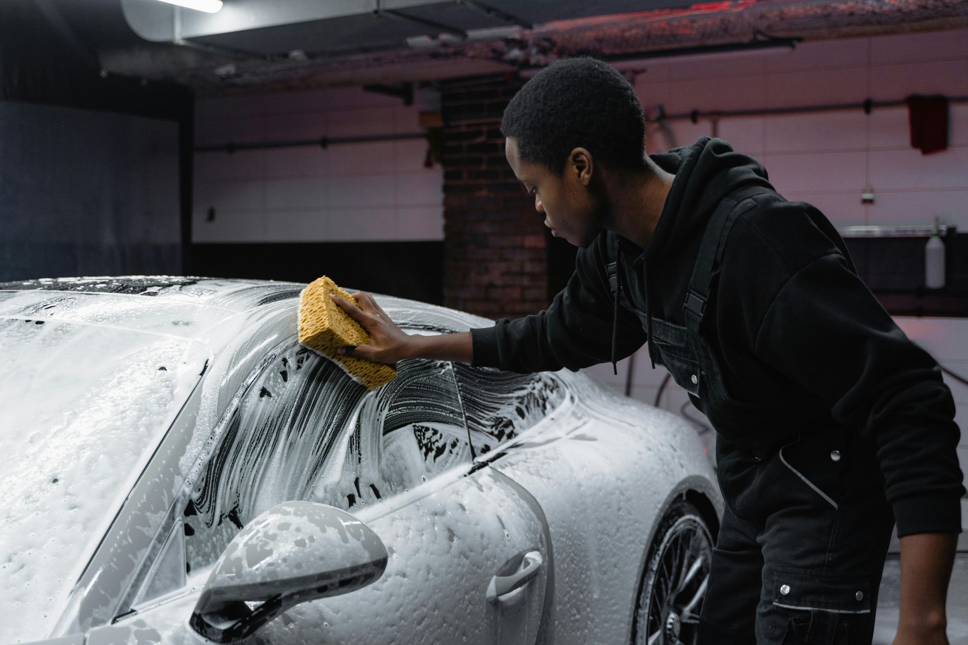 A young man washing a white car with a sponge and soapy water in a garage.