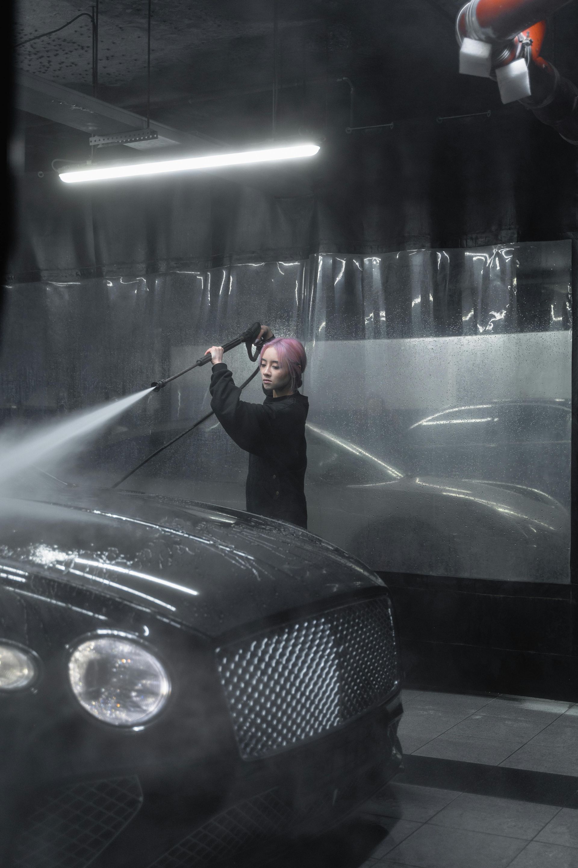 Person with pink hair washes a black car with a pressure washer in a car wash bay.
