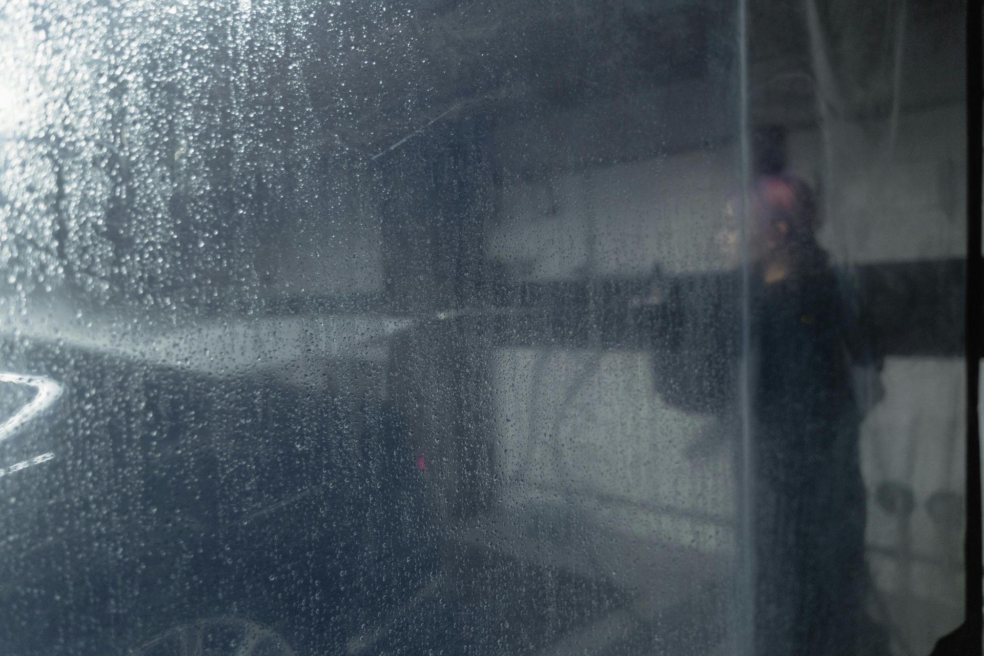 Car being washed at a car wash, viewed through a wet window with a blurry figure in the background.