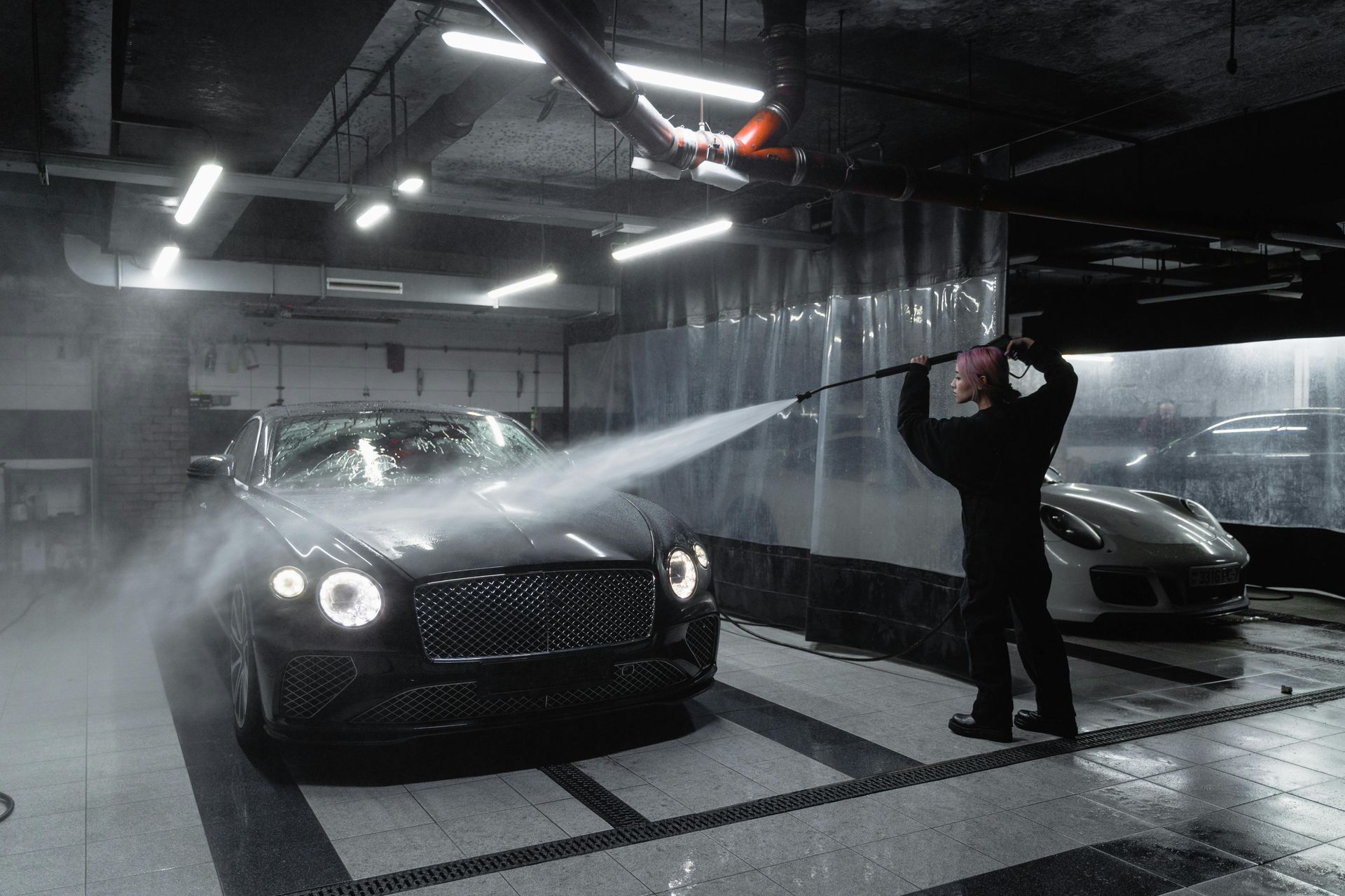 A person washing a dark car in a car wash bay; water sprays, another car visible.