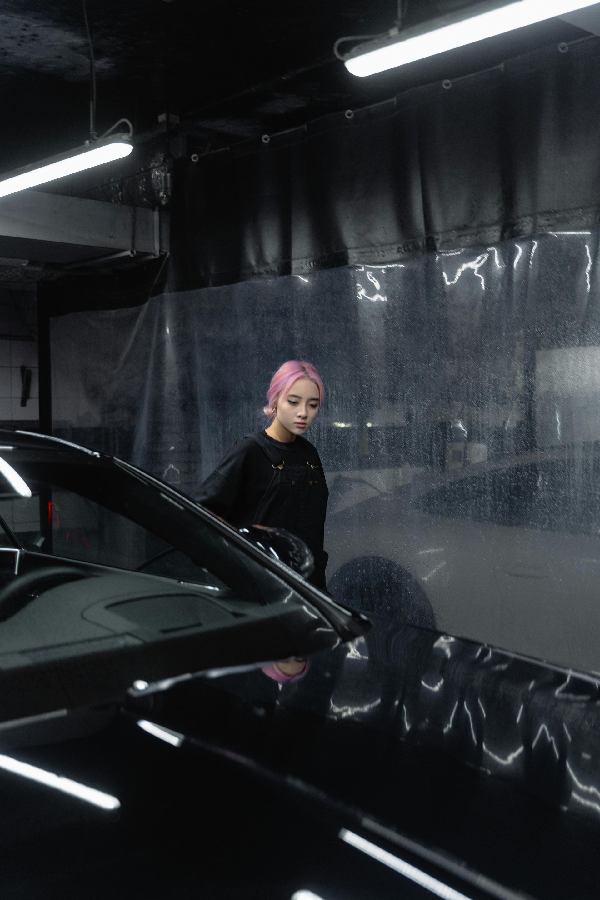 Person with pink hair washing a black car in a garage; focused expression, dark setting, reflective surface.