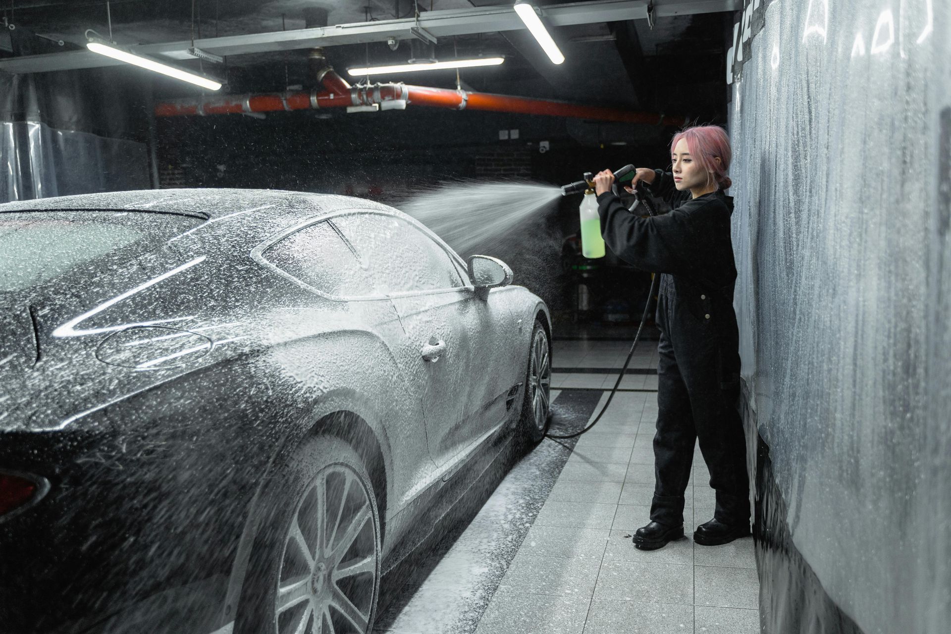 Person in black overalls sprays foam on a black car in a car wash bay.