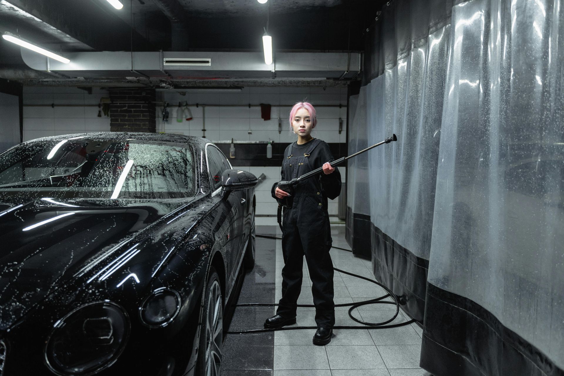 Person with pink hair washing a black car with a pressure washer in a car wash bay.