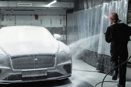 Person washing a car with foamy soap. The car is silver and in a car wash.