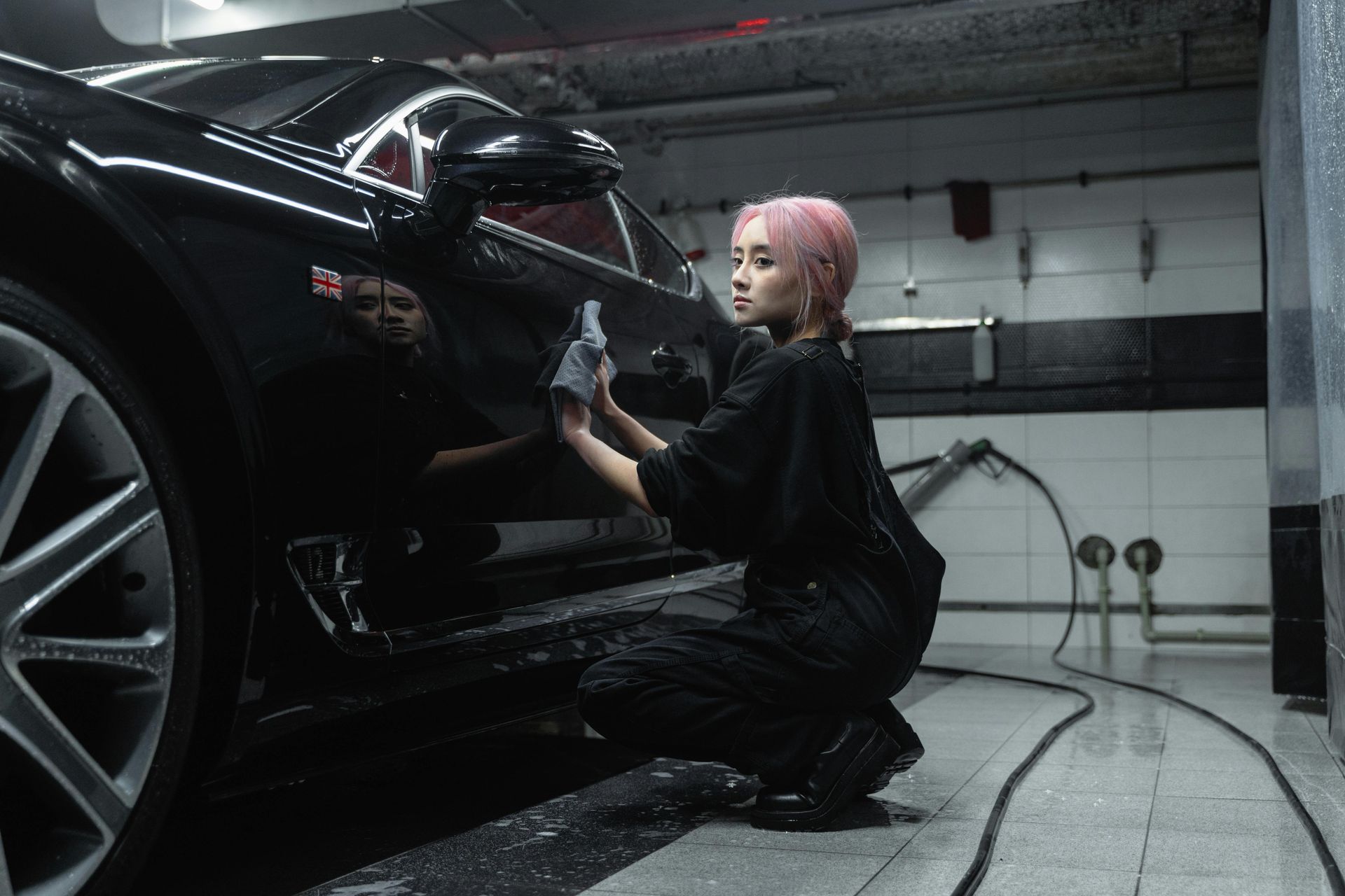 Woman with pink hair washes a black sports car in a car wash, crouching and wiping the side panel with a cloth.