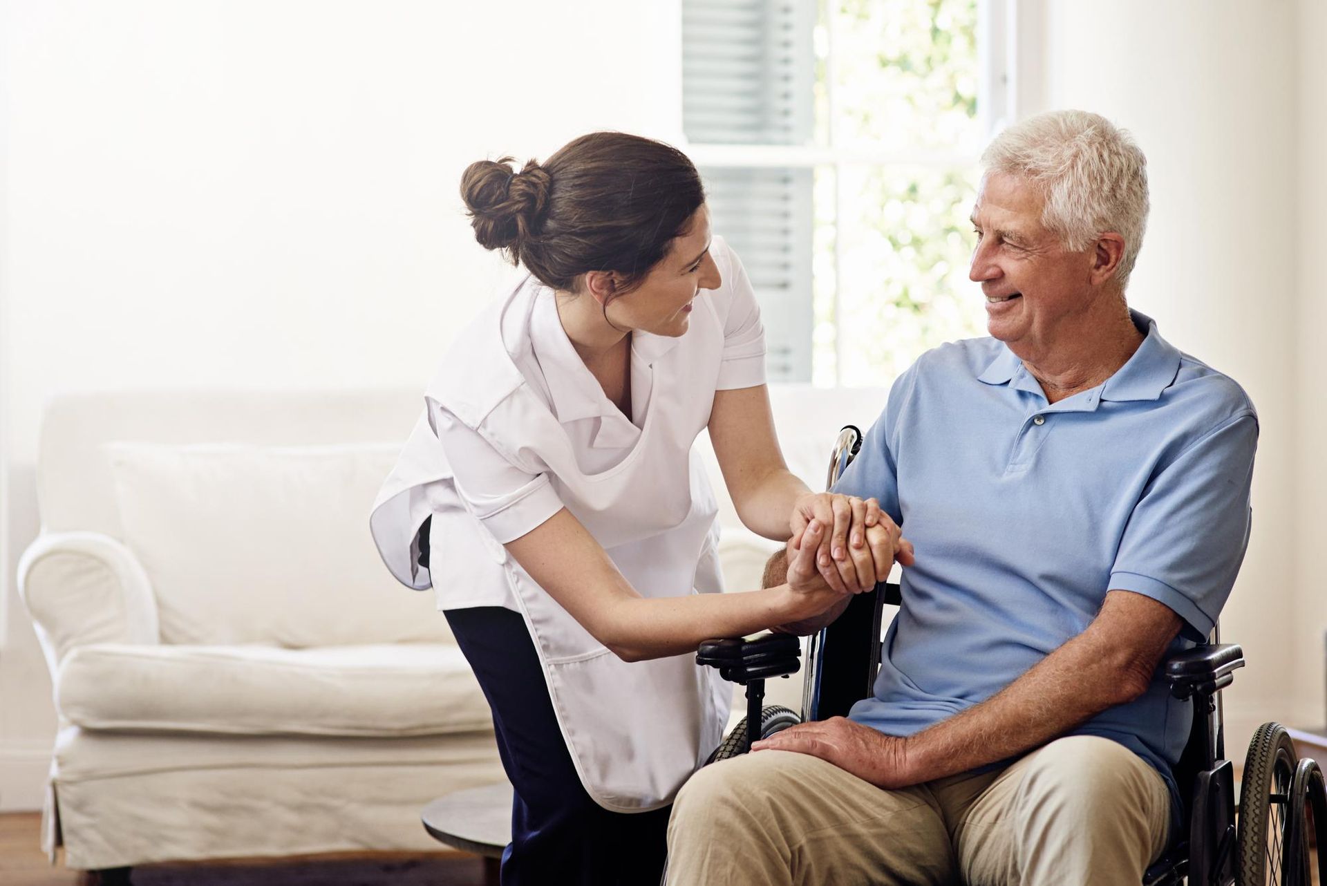 Caregiver assisting an elderly man in a wheelchair, smiling in a brightly lit room.