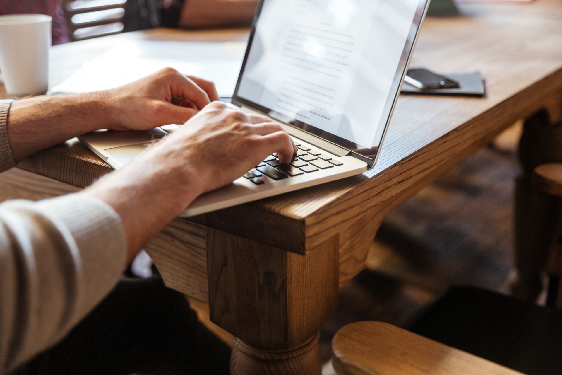 Person typing on a laptop at a wooden table, with a coffee cup and phone nearby.