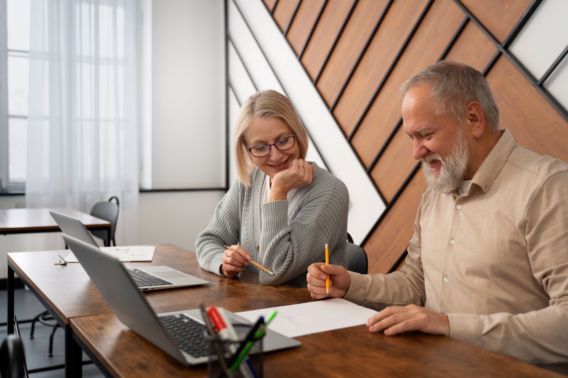 Older man and woman at a desk, smiling while working on paper. Laptops and pencils are visible.