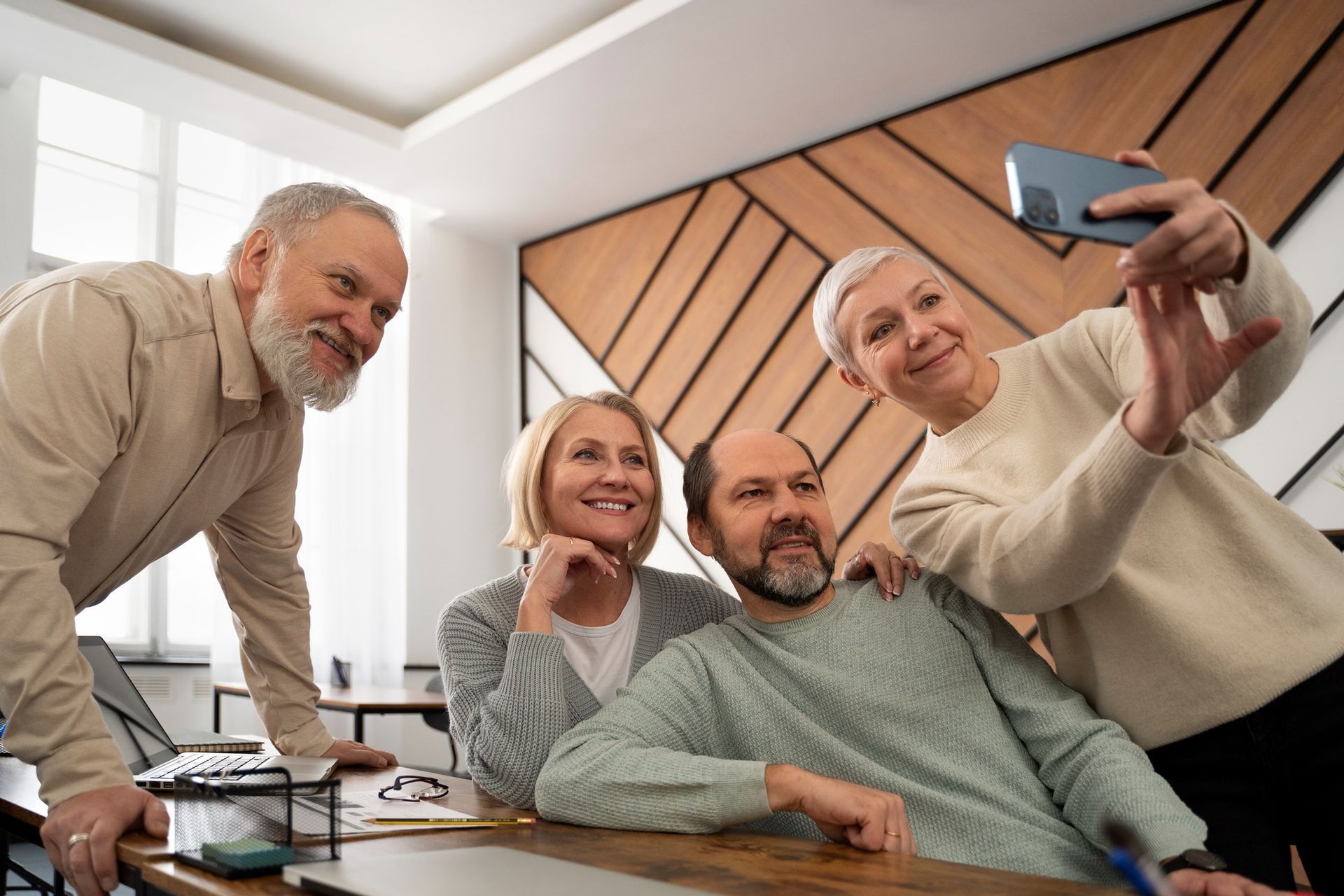 Four older adults taking a selfie in a room with a wooden accent wall; smiling.