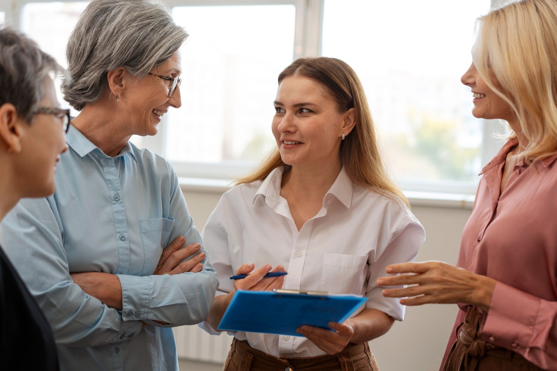 Four women in an office setting, conversing and smiling. One holds a clipboard.
