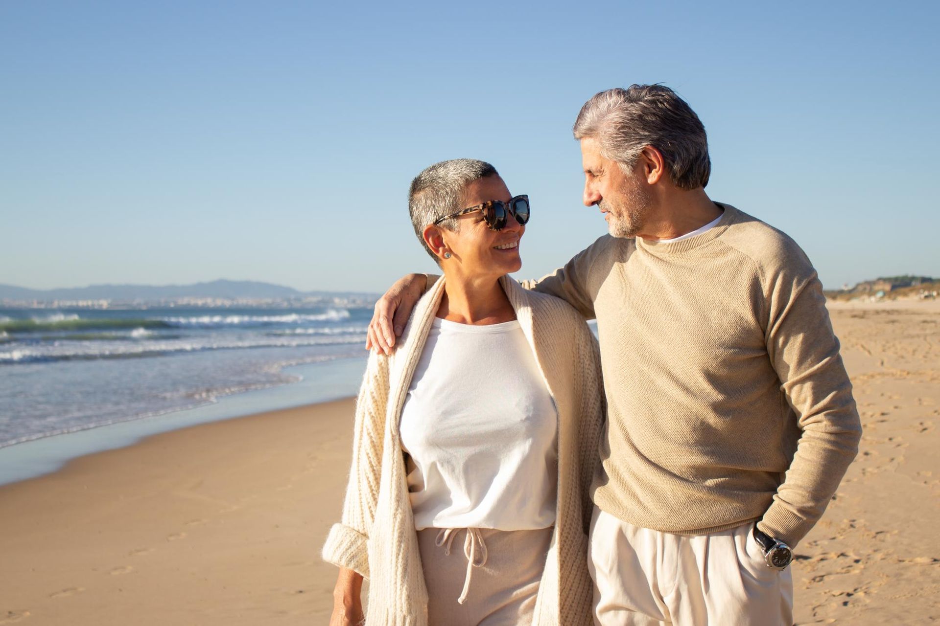 Couple walking on a sunny beach, man with arm around woman looking at each other, ocean in background.