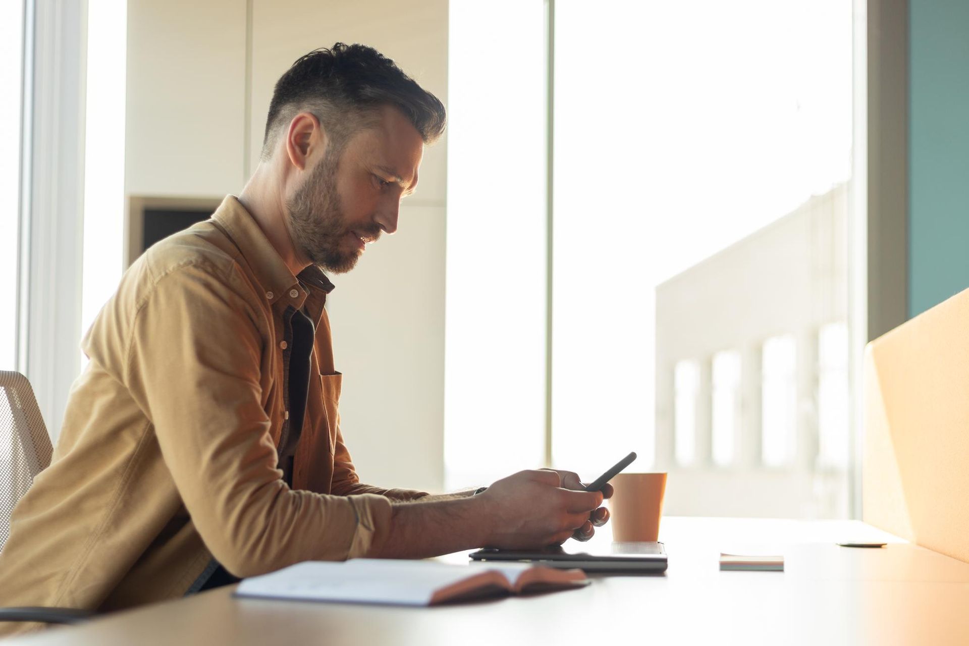 Man at desk using phone near a window. He is wearing a brown shirt. Coffee and notepad are nearby.