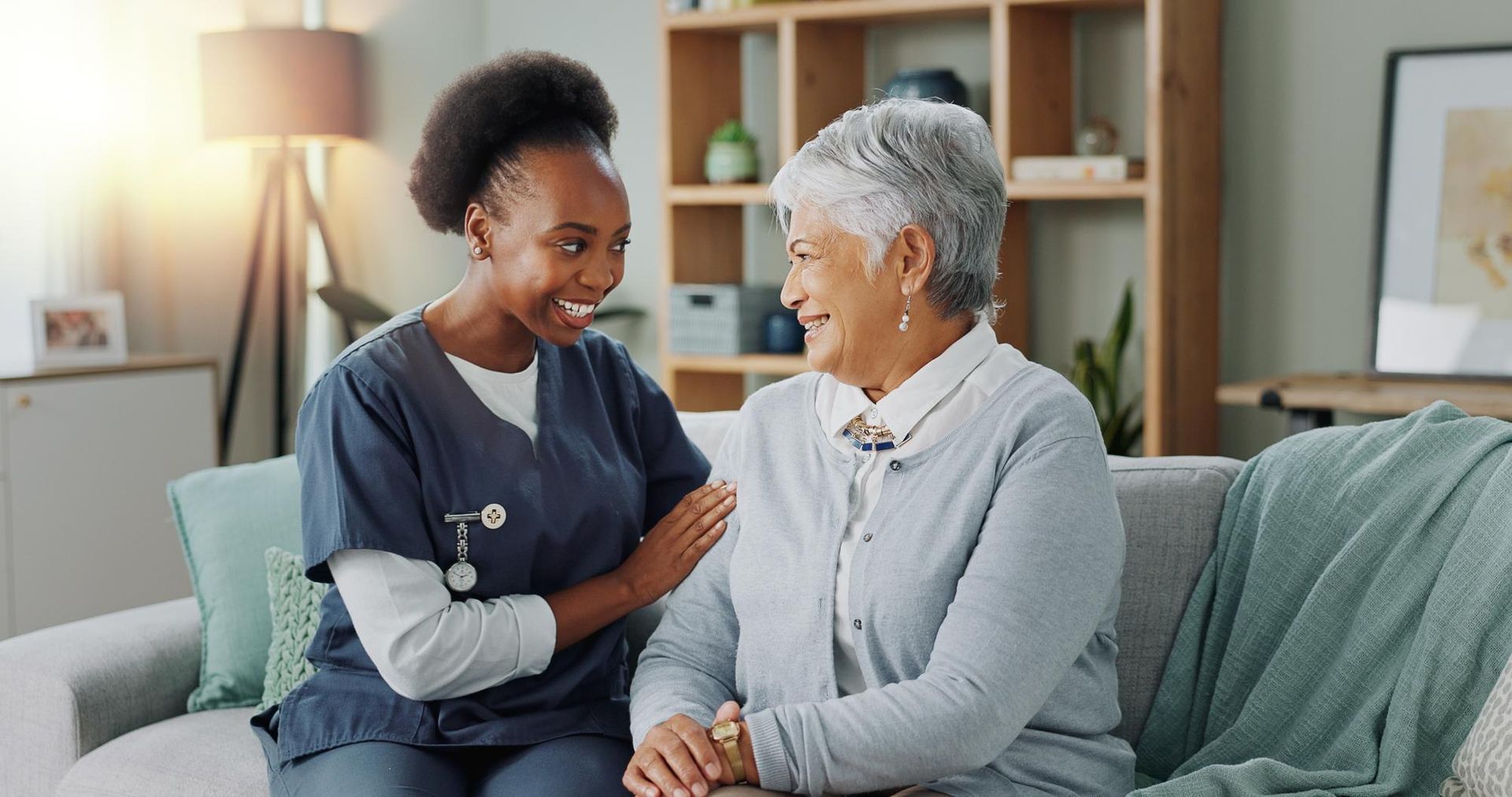 A smiling caregiver in scrubs comforts an elderly woman on a sofa.
