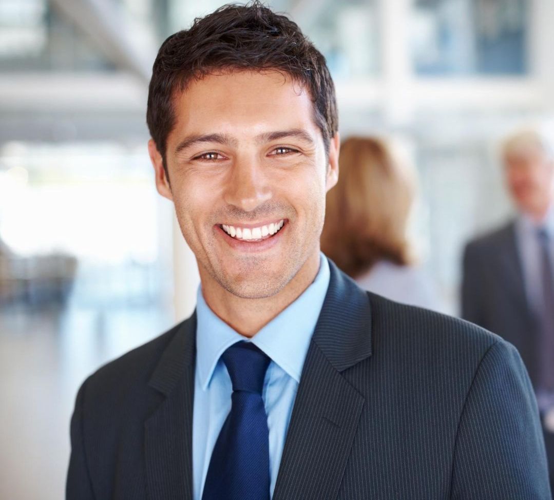 Smiling businessman in a suit and tie, light blue shirt, blurry office background.