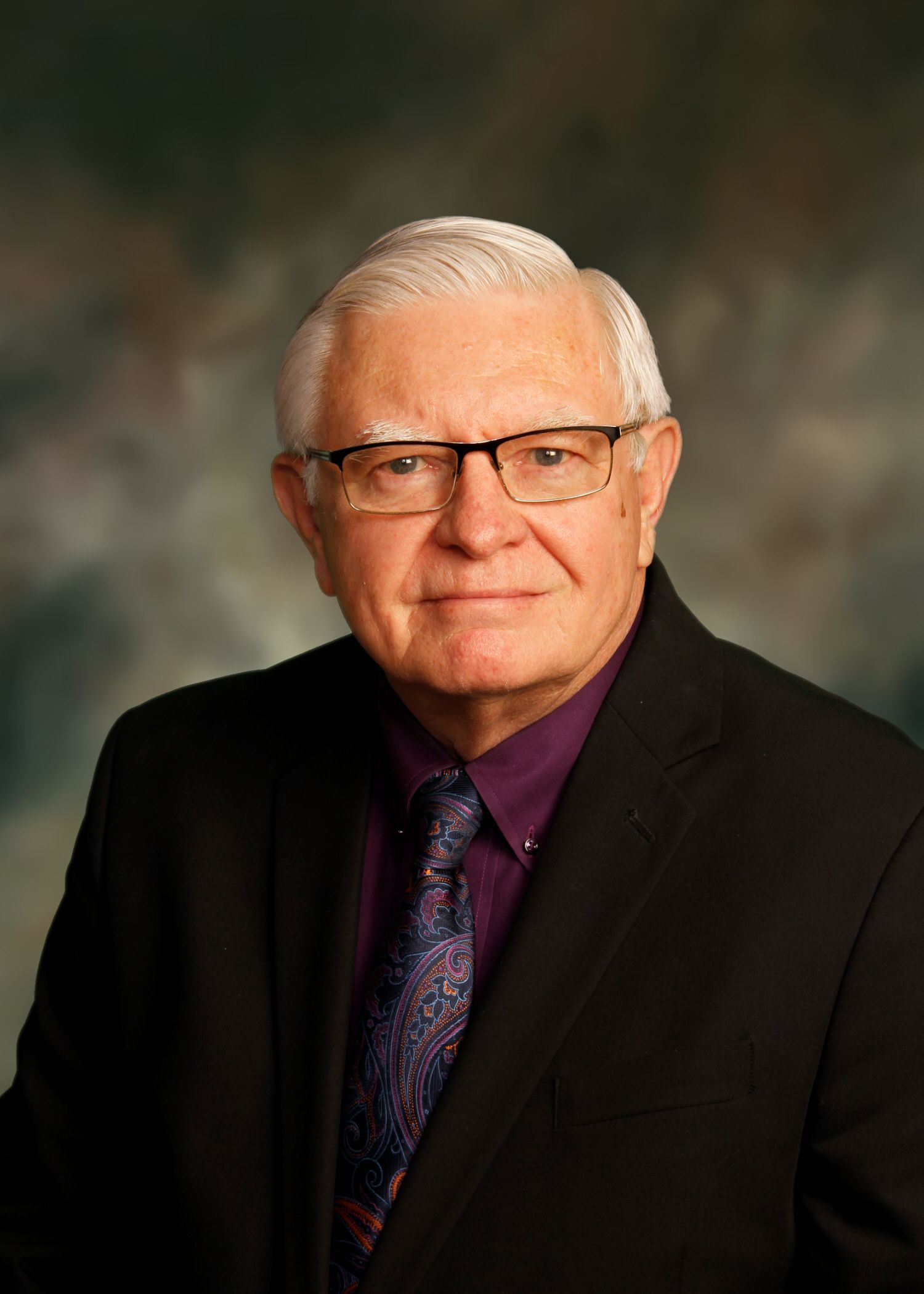 Older man wearing glasses, dark suit, purple shirt and tie.