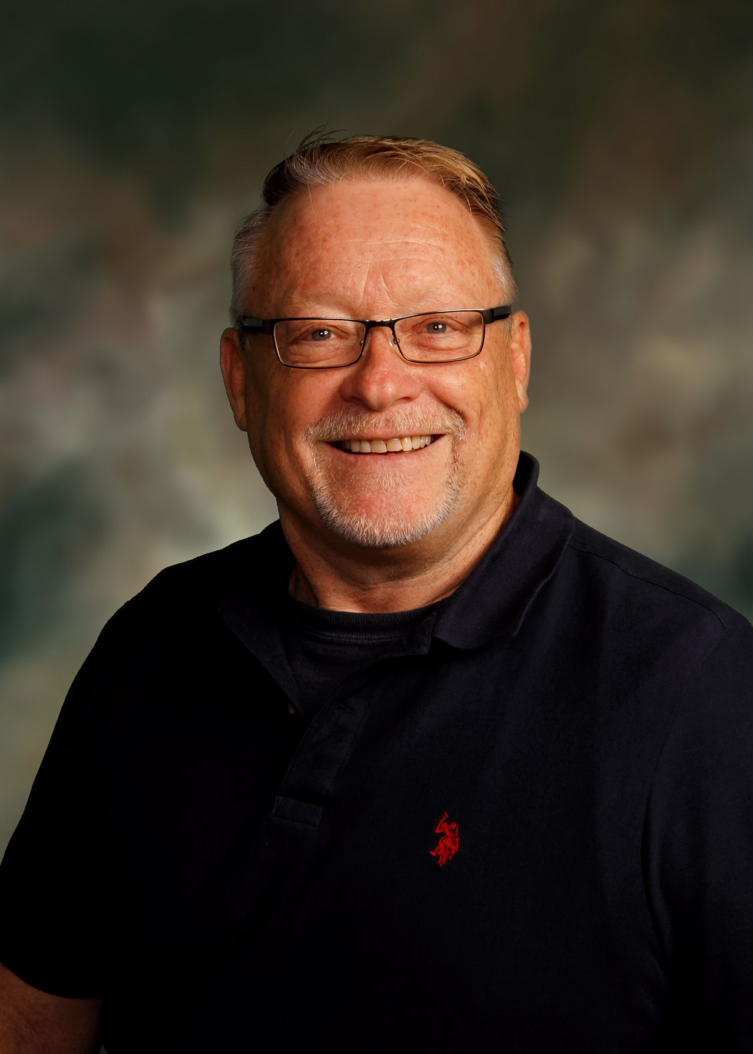 Man with glasses smiles, wearing a navy blue polo shirt, against a blurry, shaded backdrop.