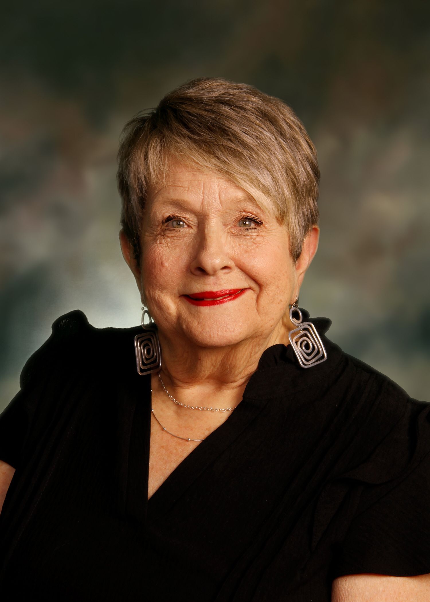 Elderly woman with short blonde hair, red lipstick, and square earrings smiles, wearing a black top.