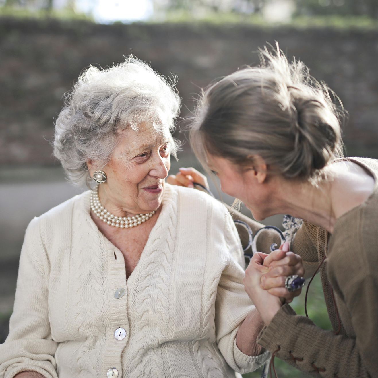 Older woman in white cardigan, pearl necklace smiles at younger woman holding her hand outdoors.