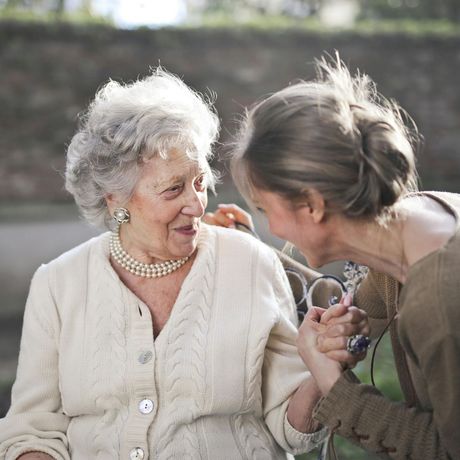 Older woman in white cardigan, pearl necklace smiles at younger woman holding her hand outdoors.