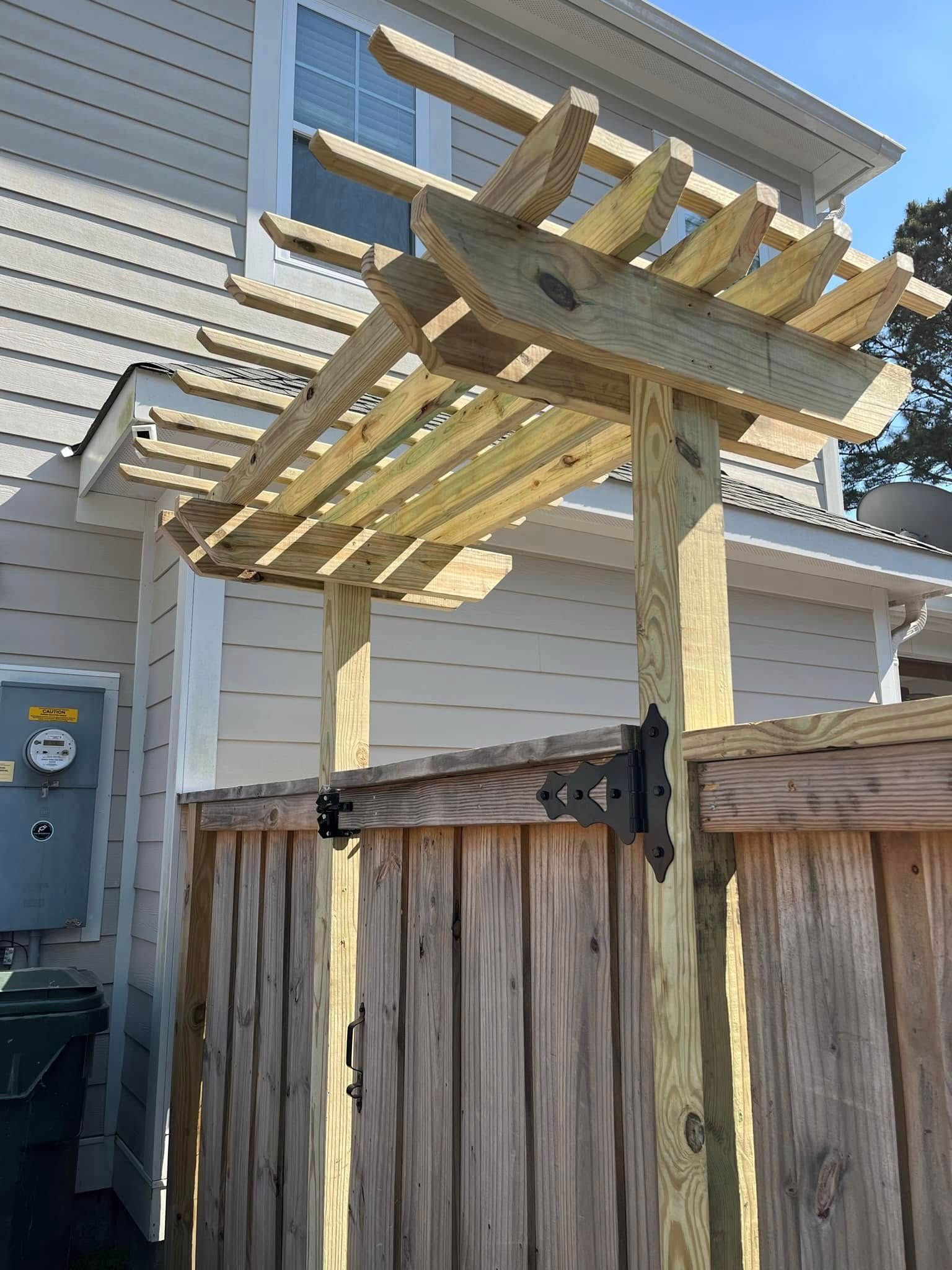 A wooden pergola is sitting on top of a wooden fence next to a house.